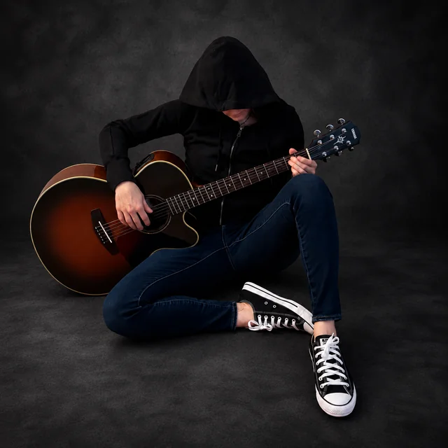 Hooded musician seated playing an acoustic guitar